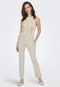 Donna che indossa un top senza maniche in maglia color crema, pantaloni beige a vita alta dritti e scarpe basse color beige chiaro, in piedi con una mano in tasca.