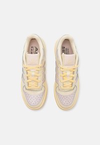 Vaaleanbeiget lenkkarit, joissa on sileä nahkapinta, purppurat yksityiskohdat, rei'itetty kärki ja keltaiset nauhat. Sivulla on kolme sinistä raitaa.