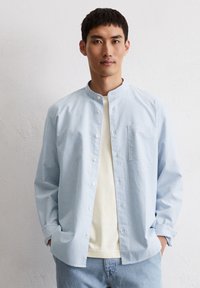 Camicia a righe blu chiaro con colletto a band, chiusura con bottoni e taschino sul petto, indossata sopra una camicia color crema, abbinata a jeans di denim chiaro.