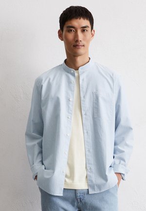 Chemise rayée bleu clair avec col bannière, devant à boutons et poche poitrine, portée sur une chemise crème, associée à un jean en denim clair.