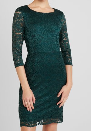 Kobieta w dopasowanej ciemnozielonej koronkowej sukience z rękawami do połowy ramienia i kwiatowym wzorem, stojąca na jednolitym szarym tle.