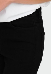 Mustad denim püksid siledat tekstuuri, millel on esipocket ja tavaline vöökoht. Asetatud valge särke taustale.