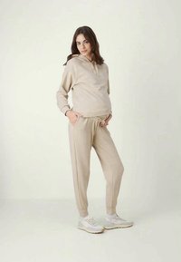 Bež dukserica i trenirke od mekog materijala, s kapuljačom na užad, elastičnim pojasom i opuštenim krojem, u kombinaciji s bijelim tenisicama.