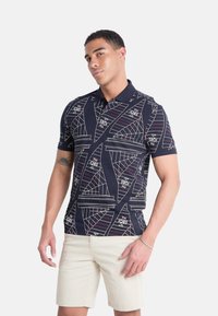 Polo de navy con patrones geométricos de veleros blancos y acentos de texto. Manga corta y cuello clásico, fabricado en una tela transpirable.