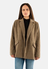 Mujer vestida con un blazer de pana marrón sobre una blusa negra y jeans azules, de pie contra un fondo blanco sencillo.