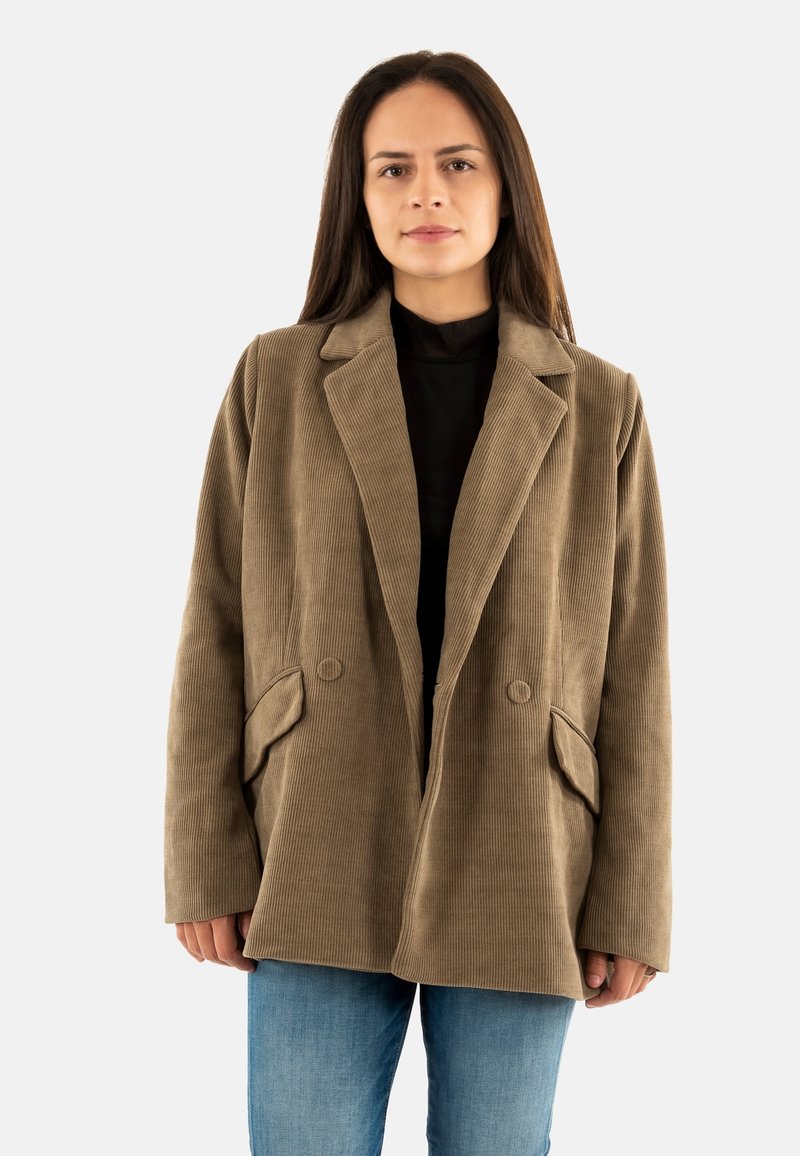 Mujer vestida con un blazer de pana marrón sobre una blusa negra y jeans azules, de pie contra un fondo blanco sencillo.