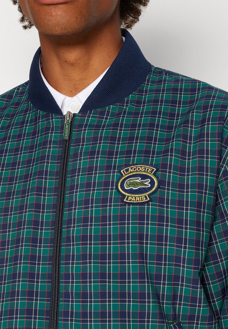 Veste bombers à carreaux en vert et marine. Dotée d'un col côtelé, d'une fermeture éclair et d'un patch logo brodé sur la poitrine gauche. Texture de tissu lisse.