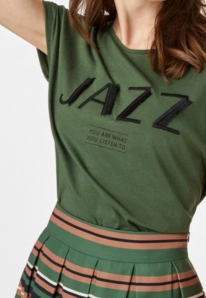 Kvinde iført en grøn t-shirt med teksten "JAZZ" og et plisseret skørt med grønne, brune, sorte og hvide striber.