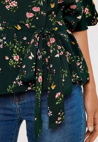 Blouse florale verte avec des fleurs roses, des papillons et un détail de taille nouée, en tissu léger, associée à un jean en denim.