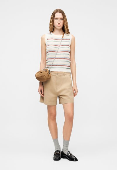 Femme portant un haut sans manches en maille rayée, un short beige, des chaussettes grises, des mocassins noirs et un petit sac bandoulière en tissage marron.