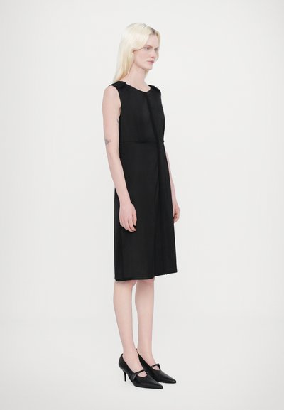 Robe noire sans manches avec une silhouette droite, longueur genou, en tissu doux, dotée d'une fermeture éclair à l'avant et de détails de couture subtils. Associée à des talons noirs.