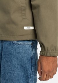 Khaki zelená bunda s elastickými manžetami, s bílou nálepkou s nápisem "Element Skateboard Co." V kombinaci s modrými denimovými džíny a texturovanou pletí.