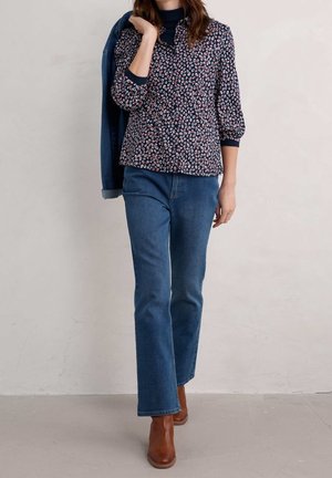 Kvinde iført blå mønstret bluse, blå jeans og brune støvler med en blå jakke over skulderen mod en ensfarvet baggrund.
