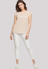 Beige short-sleeve t-shirt with a white heartbeat and mountain graphic, paired with fitted white jeans and white sneakers. Smooth texture.