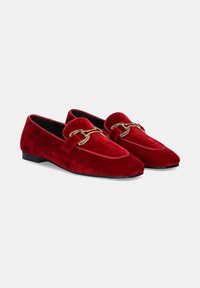 Mocasines de terciopelo rojo con suela plana negra, adornados con un detalle de cadena dorada en la parte superior. Diseño de punta afilada y textura suave.