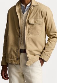 Muž v béžovém saku přes krémový svetr s pleteným vzorem a pruhovanou košili, s krémovými kalhotami a hodinkami na levém zápěstí.