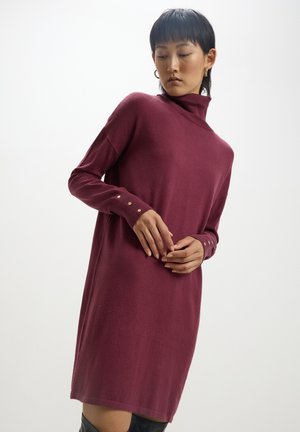 Abito a collo alto color bordeaux realizzato in materiale morbido; presenta maniche lunghe con dettagli di bottoni dorati, vestibilità rilassata e orlo al ginocchio.