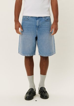 Hellblaue Jeansshorts mit entspannter Passform, ausgestattet mit fünf Taschen, gesteppten Säumen und einem Knopfverschluss. Getragen mit grauen Socken und schwarzen Schuhen.