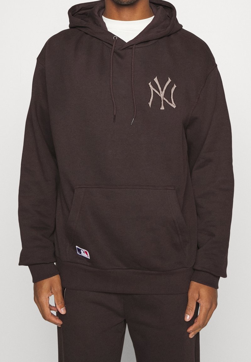 Homme portant un sweat à capuche marron foncé avec le logo des New York Yankees sur la poitrine et le logo MLB près de la poche, assorti d’un pantalon assorti.