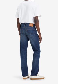 Blå denimjeans med rak passform, försedda med en tan läderlapp på den bakre midjan och en klassisk femficksdesign.