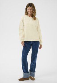 Pull crème avec détails de broderie florale aux épaules, associé à un jean bleu à jambes larges avec un ourlet roulé. Des chaussures marron complètent le look.