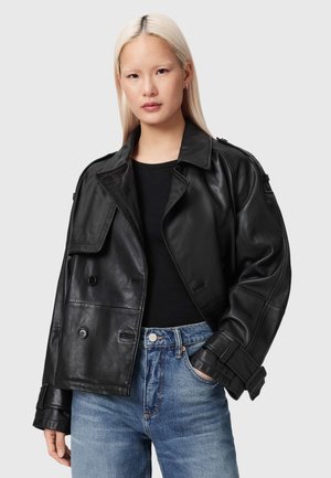 Jeune femme aux longs cheveux blonds portant une veste en cuir noire, un haut noir et un jean bleu, debout la main dans la poche.