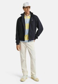 Marine zip-up jack over een gestreepte polo in blauw en geel, crèmekleurige broek en lichtgekleurde sneakers, met een witte pet met blauw logo.