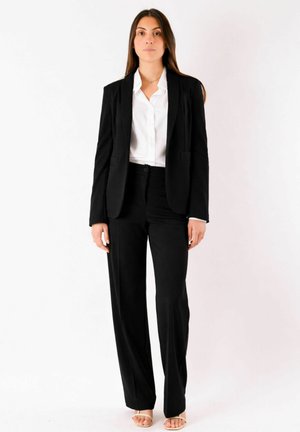 Donna con capelli castani lunghi che indossa un completo nero con pantaloni a gamba larga e una camicetta bianca, in piedi davanti a uno sfondo bianco semplice.