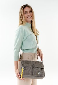 Donna sorridente con maglione verde chiaro e pantaloni beige che tiene una borsa a tracolla grigia con due tasche frontali e zip gialla.