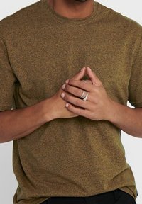 Kortärmad t-shirt med rund halsringning i ett strukturerat olivgrönt tyg, med ett subtilt prickigt mönster. Händerna är i kors framför kroppen och det bärs ringar.