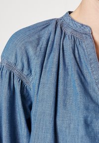 Top in denim con scollatura rotonda, dettagli arricciati e accenti di cucitura sottili. Tessuto blu chiaro con una texture leggera.