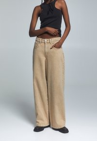 Beżowe dżinsy o szerokich nogawkach z wysokim stanem, posiadające pięć kieszeni, zapięcie na guzik oraz wyblakłą fakturę materiału dżinsowego. Ubrane z czarnym topem bez rękawów.