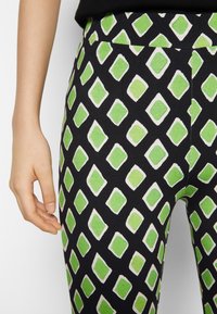 Legginsy na czarnym tle z powtarzającym się wzorem zielonych diamentów obramowanych na biało, noszone z czarną bluzką.