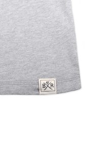 Grå bomulds t-shirt kant med et lille, firkantet vævet mærke med sort tekst og krydsede grafikker. Blød tekstur med en ren finish.