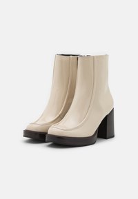 Botines de charol beige hasta el tobillo con punta cuadrada, detalles de costura elevada y un tacón de madera grueso. Textura suave, diseño minimalista.