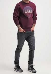 Sudadera burdeos con gráfico "CORE", jeans ajustados gris, y zapatillas negras/grises con estampado. Presenta puños y cuello acanalados.