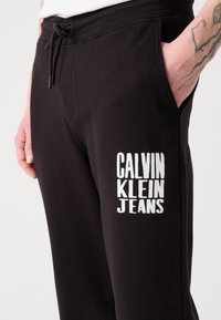 Melni kokvilnas sporta bikses ar elastīgu jostasvietu un auklu. Uz kreisā aug�šstilba ir balta izšūta logo "CALVIN KLEIN JEANS".