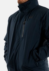 Veste imperméable navy avec col haut, poche poitrine zippée et poignets réglables. Texture lisse avec un design épuré et minimal.