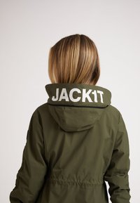 Person med blondt hår if�ørt en olivengrøn jakke med høj krave, der viser teksten "JACK1T" i hvide bogstaver på ryggen.