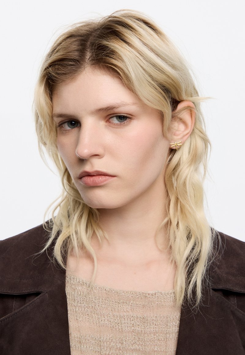 Pendientes de oro con un diseño geométrico, suéter ligero texturizado y chaqueta de ante marrón. El cabello rubio largo y ondulado enmarca la cara.