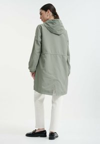 Khaki zelená nepremokavá bunda s kapucňou, bočnými rozparkami, elastickým pásom a dlhými rukávmi. Nosená cez krémové nohavice a čierne topánky.