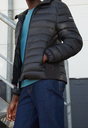 Sort puffjakke med hætte, prydet med et quiltet design og lynlåslommer, båret over en blå skjorte og suppleret med mørke jeans.