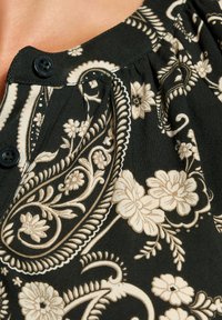 Nahaufnahme eines schwarzen Stoffs mit beigen Blumen- und Paisleymustern, der zwei schwarze Knöpfe in der Nähe eines gerafften Ausschnitts zeigt.
