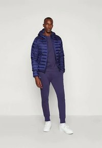 Casaco acolchoado azul-marinho com costuras horizontais, combinado com um sweatshirt e calças de treino em correspondência. Ténis brancos completam o look.