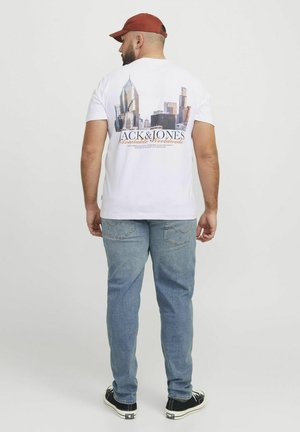 Weißes Baumwoll-T-Shirt mit einem Stadt-Skyline-Graphic auf der Rückseite in Blau- und Grautönen, kombiniert mit hellblauen Jeans und schwarzen Sneakers.