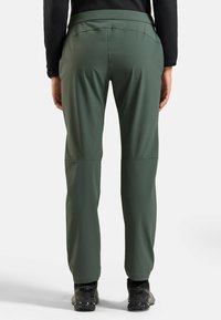 Grüne, elastische Outdoor-Hosen mit glatter Textur, schmal zulaufenden Beinen und hohem Bund. Getragen mit schwarzen Wanderschuhen.