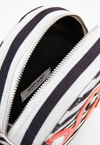 Raita kangasta valmistettu laukku, jossa on mustavalkoisia vaakaraitoja, vetoketjukiinnitys ja pieni sisätasku. Sisältää oransseja yksityiskohtia ja etiketin.