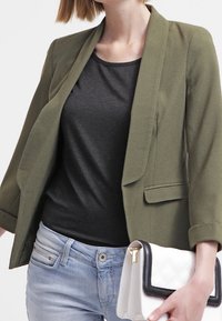 Olivově zelený blejzr s elegantním designem, který má jednu kapsu a tříčtvrteční rukávy. Slučuje se s černým topem a bílou prošívanou kabelkou.