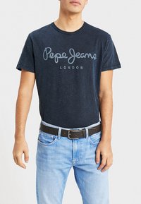 Sort bomulds t-shirt med "Pepe Jeans LONDON" logo i skråskrift, parret med lyseblå jeans og et sort bælte med sølvdetaljer.