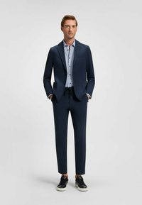 Completo blu navy con giacca aderente, abbinato a una camicia azzurra con pois chiari e pantaloni affusolati, completato da sneaker nere.